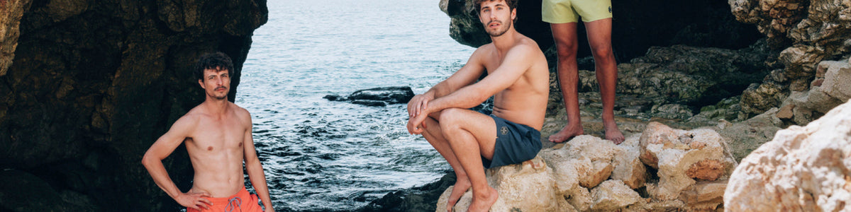 Zwei Männer in Badeshorts auf Felsen am Meer.