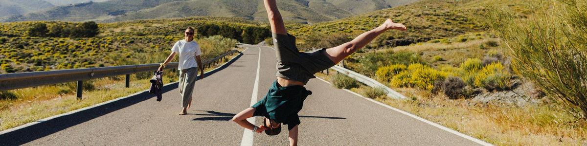 Frau geht eine Landstraße entlang, daneben macht ein Mann einen Handstand.