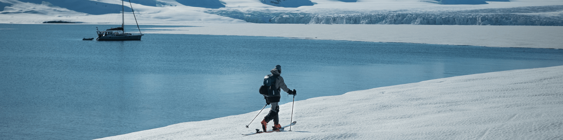 In diesem Bild sieht man einen Mann, der eine Schneetour entlang eines Sees macht. Auf dem See ist ein Boot zu sehen. Der Hintergrund zeigt eine eisige Schneelandschaft.