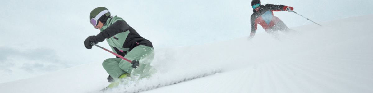 Zwei Skifahrer fahren mit Schneefahne einen Pistenhang hinunter
