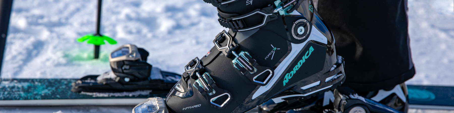 Skischuh von Nordica in schwarz/türkis/weiß zu erkennen. Dieser wird in einen Skier eingeschnallt. Im Hintergrund ist Schnee zu erkennen.