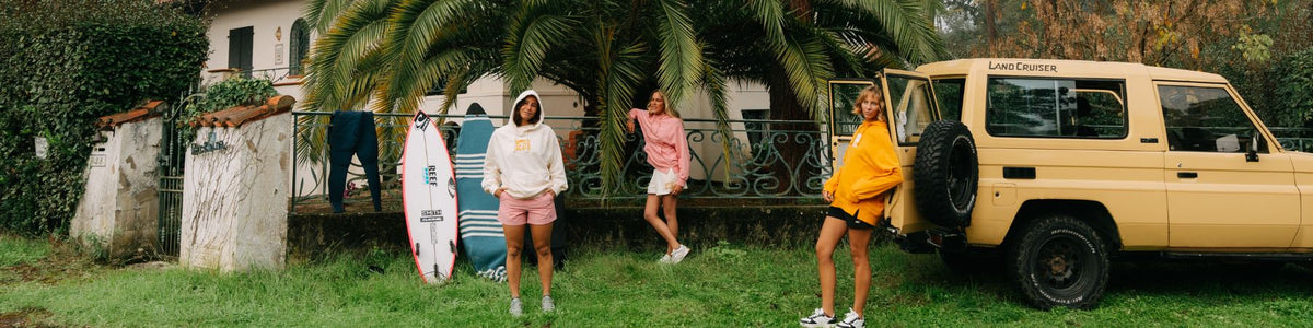 Drei Frauen in Pullovern und Hoodies vor einer Mauer, daneben ein Geländewagen und ein Surfbrett.