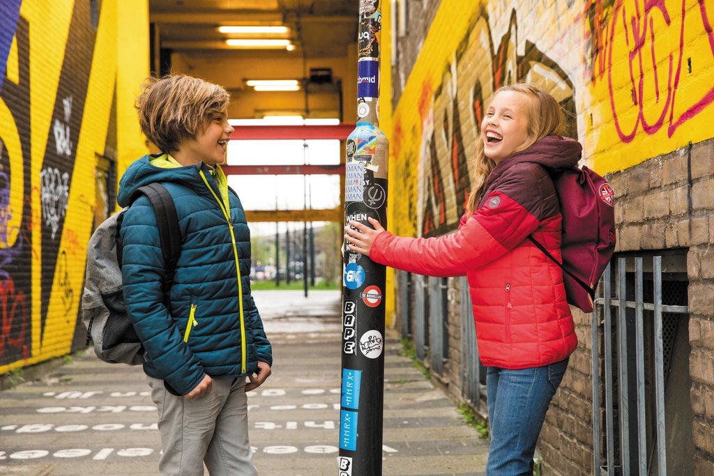 Zwei Kinder in Steppjacken mit Rucksack vor einer Graffitiwand