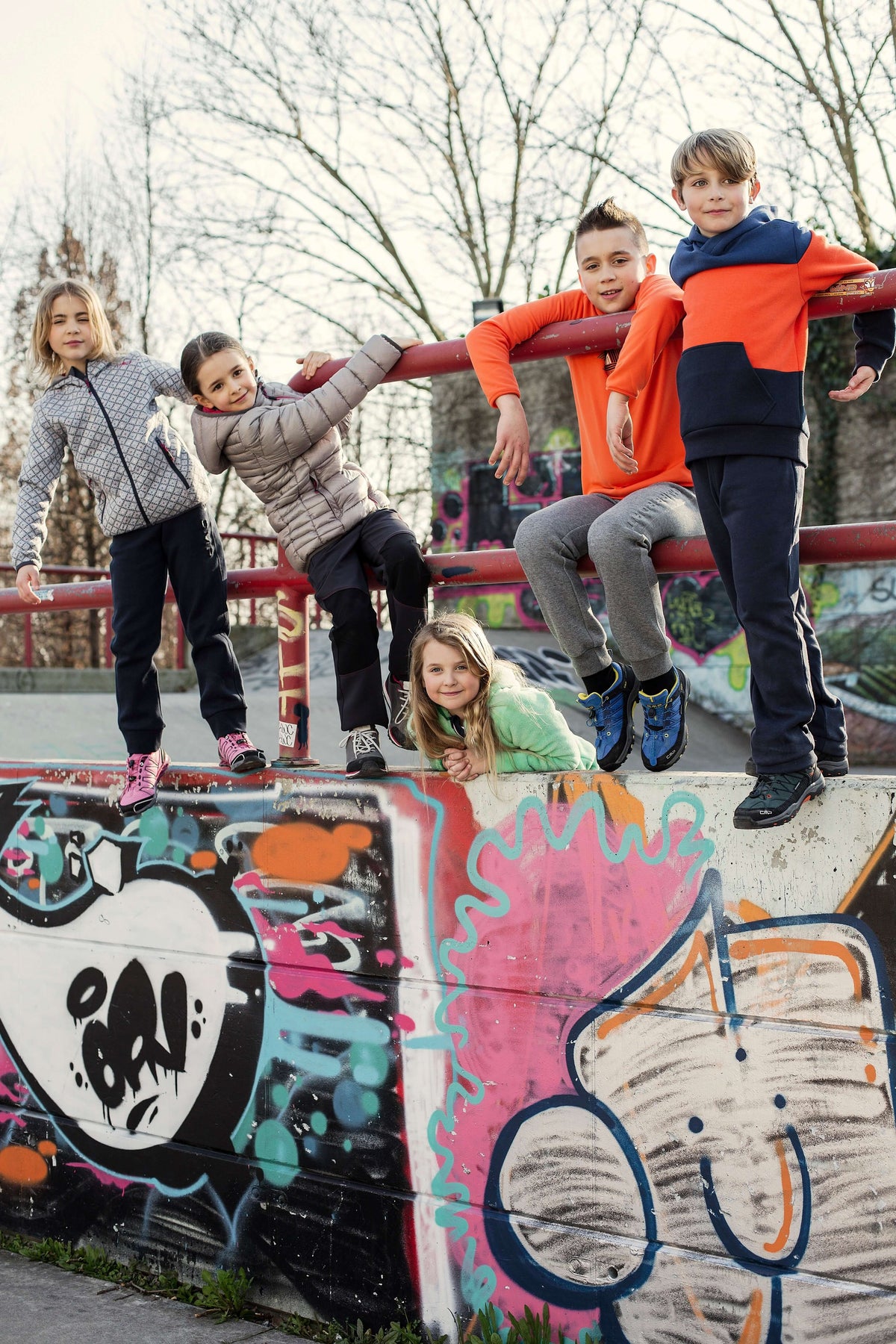 Fünf Kinder in Outdoorjacken sitzen auf einem Geländer über einer Graffitiwand