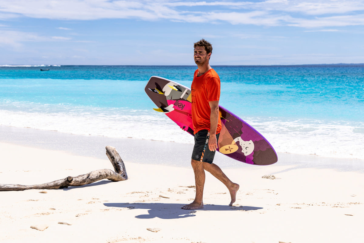 Mann in schwarzer Badeshorts und orangenem Freizeitshirt läuft Surfbrett in der Hand vom Strand.